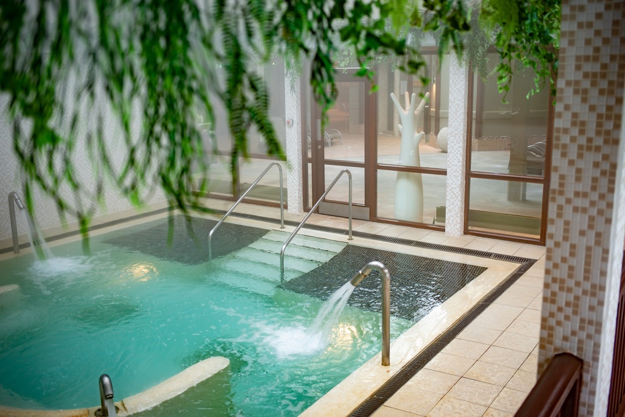 Spa pool bubbles and cascades, water jets pouring from stainless spouts and steps descending between handrails. Surrounded by tiled flooring, glass walls, and hanging greenery in a calm, indoor wellness area.